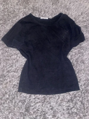 Svart frotté t-shirt - En enkel svart tajt t-shirt i mjukt frottématerial. T-shirten har rund halsringning och korta ärmar, perfekt för en avslappnad och bekväm stil. Passformen är normal och tyget ger en lite sportig känsla.
