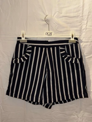 Mötkblå shorts med vita ränder - Snygga mörkblåa shorts med vita vertikala ränder och dekorativa fickor framtill. Shortsen har en relaxed passform och är perfekta för varma dagar. Stilsäkert val för dig som gillar mönster och vill sticka ut lite extra. 