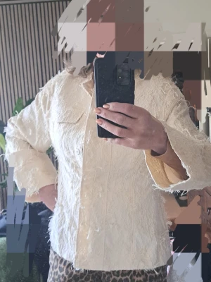 Offwhite jack med fransar, L - Säljer en offwhite overshirt med fransiga detaljer och knappar framtill. Jackan har en avslappnad passform, bröstficka och är långärmad. Perfekt för dig som vill sticka ut med en unik och trendig look.
