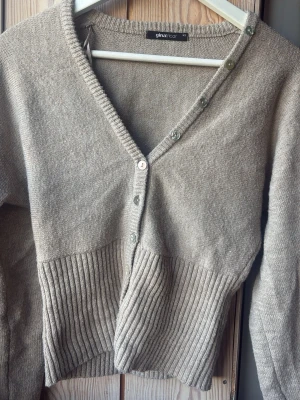 Beige ribbad kofta från Gina Tricot - Snygg beige kofta från Gina Tricot med v-ringning och knappar längs hela framsidan. Koftan har ribbad nederdel och långa ärmar, vilket ger en trendig och avslappnad look. Perfekt att slänga över en topp för extra stilpoäng.