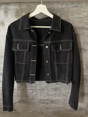 Svart croppad jeansjacka  - Svart jeansjacka med kontrasterande vita sömmar och fransad nederkant. Jackan har två stora bröstfickor och silvriga knappar.
