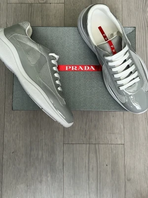 Grå sneakers från Prada - Snygga grå sneakers från Prada med vita detaljer och vita skosnören. Skorna har en kombination av glansigt material och mesh för en modern look. Sulan är vit och ger en sportig känsla. Perfekta för dig som vill ha stilrena och trendiga sneakers.