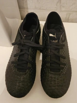 Svarta fotbollsskor från Puma - Säljer ett par svarta fotbollsskor från Puma med klassisk snörning och strukturmönstrad ovandel för bättre bollkontroll. Skorna har låg profil och dobbar för spel på gräs. Snyggt Puma-logga på sidan och tungan. Perfekta för dig som vill ha stilrena och funktionella skor på planen.