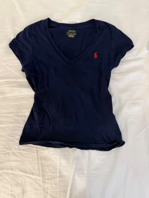Marinblå v-ringad t-shirt Polo Ralph Lauren - Snygg marinblå t-shirt från Polo Ralph Lauren med v-ringning och klassisk röd logga broderad på bröstet. T-shirten har korta ärmar och är gjord i mjuk bomull som känns skön mot huden. Perfekt för en clean och stilren look❤️