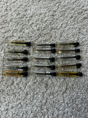 Essnce parfymer - Säljer ett set med 12 parfymprover i små genomskinliga glasflaskor med svart kork. Varje flaska har tryckt text med olika doftnamn som Irresistible Enigma, Milky Flower, Iconic Flower och fler. Perfekt för dig som vill testa olika dofter innan du bestämmer dig.