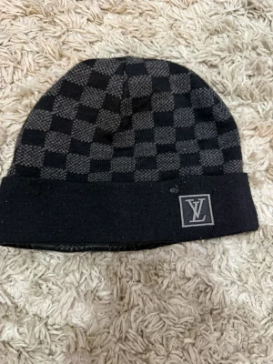 Svart och grå Louis Vuitton-mössa - Snygg svart och grå mössa från Louis Vuitton med klassiskt rutmönster och vikt kant. LV-logga framtill. Perfekt accessoar för att hålla värmen och samtidigt se trendig ut. Passar till de flesta outfits och är enkel att matcha.
