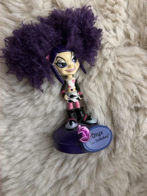 Onyx Trollz figur med lila hår - Cool samlarfigur av Onyx från Trollz med stort fluffigt lila hår, svart och rosa outfit samt glittriga skor. Figuren står på en lila bas med namnplakett. Perfekt för dig som gillar unika prydnader eller vill ha nostalgi från Trollz.