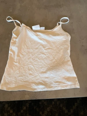 Vit enkel linnetopp med smala band - Säljer en vit linnetopp med smala axelband. Toppen har en rak passform och är tillverkad i mjuk bomull, perfekt för varma dagar. Enkel och stilren design som funkar till det mesta. Fräsch och lätt att matcha med olika outfits.