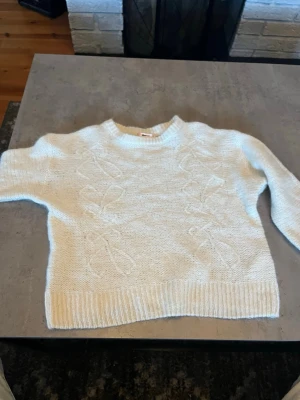Vit kabelstickad tröja - Mysig vit stickad tröja med kabelstickat mönster framtill. Tröjan har rund halsringning, långa ärmar och ribbade muddar vid ärmslut och nederkant. Perfekt för kyliga dagar och enkel att matcha med jeans eller kjol. Den är liten i storleken passar 140!