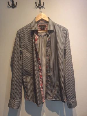 Randig skjorta från Swedish Shirt Design - Snygg svartvit randig skjorta från Swedish Shirt Design med slim fit. Insidan har ett rutigt mönster i rött, vitt och grått. Klassisk krage och långa ärmar. Perfekt för dig som gillar stilrena detaljer.