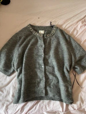 Grå kofta med diamant broderad krage - Mysig grå kofta med korta ärmar och knappar framtill. Kragen är dekorerad med diamanter och pärlor som ger en extra touch. Perfekt att slänga över en topp för en trendig look. Materialet känns mjukt och fluffigt.