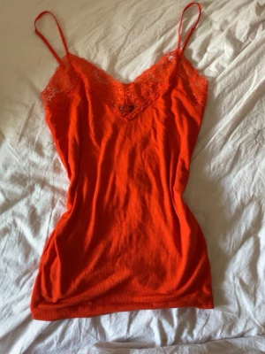Orange nattlinne med spetsdetaljer - Ett orange linnemed tunna axelband och v-ringning dekorerad med spets. Materialet är ribbat och ger en mjuk känsla mot huden. Spetsen framtill ger en extra feminin touch och linnet har figurnära passform.