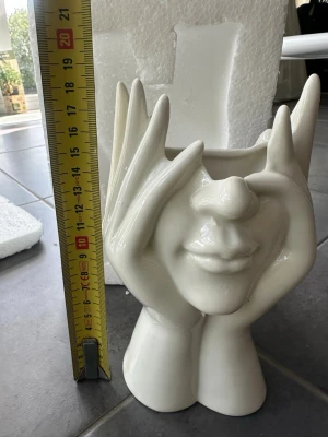 Vit keramikskulptur ansikte & händer - Unik vit keramikskulptur som föreställer ett ansikte inramat av två händer. Skulpturen är cirka 9 cm bred och har en blank finish. Perfekt som dekoration i hemmet eller som en cool inredningsdetalj på skrivbordet.