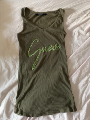 Olivgrön ribbad linnetopp Guess - Snygg olivgrön ribbad linnetopp från Guess med v-ringning och ärmlös design. Guess-loggan framtill är dekorerad med gröna paljetter som ger toppen en unik touch. Perfekt för dig som vill sticka ut lite extra.