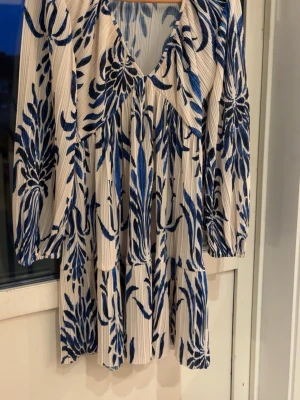 Vit och blå blommig kortklänning - Säljer en luftig kortklänning med v-ringning och långa ärmar. Klänningen är vit med blått blommönster och har en avslappnad passform. Materialet är lätt och känns skönt mot huden, perfekt för varmare dagar.