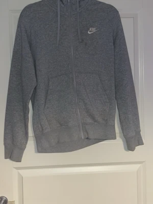 Grå zip hoodie från Nike - Säljer en grå zip hoodie från Nike med huva och dragsko. Hoodien har två fickor framtill, ribbade muddar vid ärmslut och nederkant samt en liten Nike-logga på bröstet. Perfekt för en avslappnad och sportig stil. Smuts på loggan och lite under den, inget super märkvärdigt 