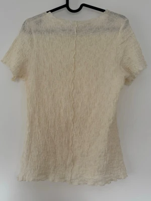 Ribbad topp med vågig söm - Trendig beige topp med ribbad struktur och vågig söm framtill och baktill. Kortärmad modell med lätt transparent material och hög hals. Står ej storlek men passar som Xs/S