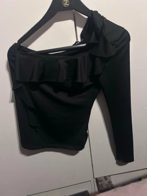 Svart oneshoulder topp från Bubbleroom - Snygg svart oneshoulder topp från Bubbleroom i stretchigt material. Toppen har en volangdetalj längs ringningen och en lång ärm på ena sidan. Perfekt för dig som vill sticka ut med en unik och trendig look.