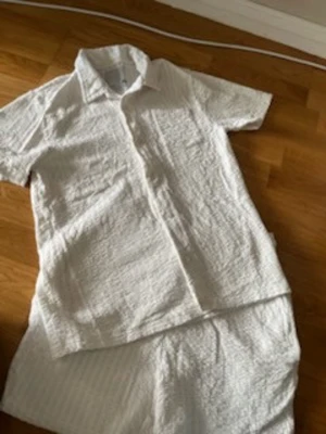 Vitt set med skjorta och shorts - Säljer ett vitt set med kortärmad skjorta och matchande shorts. Setet är gjort i ett krinklat, lätt tyg som känns luftigt och bekvämt. Skjortan har klassisk krage och knäppning framtill. Perfekt för en avslappnad och stilren look.