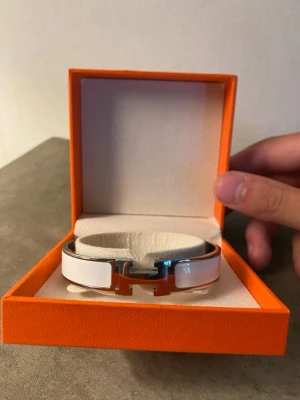 Vitt  H armband - Säljer ett stilrent vitt  Clic H armband i metall med emaljdetaljer. Armbandet har den ikoniska H-loggan framtill och kommer i original orange ask. Perfekt accessoar för att lyfta din outfit med en lyxig touch.