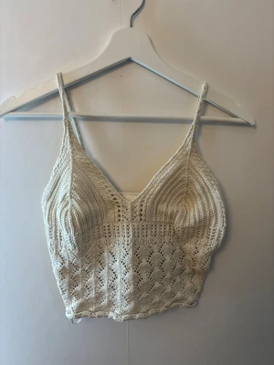 Vit virkad crop top - Säljer en söt virkad crop top i offwhite med tunna axelband och v-ringning. Toppen har ett detaljerat hålmönster och är perfekt för sommaren. Materialet är mjukt och följsamt, och modellen är kort och figurnära.