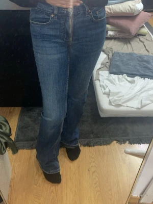 Blå bootcut jeans med hög midja - Säljer ett par klassiska blå bootcut jeans med hög midja och fem fickor. Jeansen har en snygg tvätt och subtila slitningar för en avslappnad look. Passar perfekt till sneakers eller boots och ger en tidlös stil.