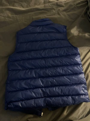 Blå dunväst från Moncler - Snygg blå dunväst från Moncler med quiltad design och hög krage. Västen har två dragkedjeförsedda fickor framtill och Moncler-logga på bröstet. Perfekt att slänga över en hoodie eller tröja för extra värme och stil.