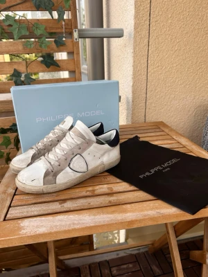 Philippe Model vita låga sneakers - Snygga vita sneakers från Philippe Model med gråa och svarta detaljer. Klassisk låg modell med rund tå, vita skosnören och ikonisk sköldlogga på sidan. Ovandelen är i läder och mocka, sulan är i gummi. Perfekta för en trendig och avslappnad stil.