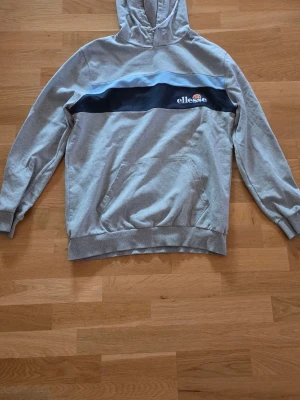 Grå hoodie från Ellesse med logga - Snygg grå hoodie från Ellesse med svart band över bröstet och broderad logga i vitt, orange och rött. Klassisk huva och känguruficka framtill. Ribbstickade muddar vid ärmar och nederkant. 