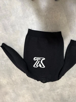 Svart stickad tröja med vit logga - Snygg svart stickad tröja med stor vit logga framtill. Tröjan har rund halsringning, långa ärmar och ribbade muddar vid ärmslut och nederkant. Perfekt för dig som gillar enkel men trendig stil.