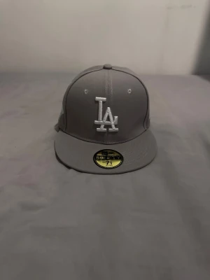 Grå keps med broderad logga - Säljer en grå snapback-keps med logga broderad framtill och en cool detalj på sidan. Kepsen har platt skärm och klassisk 59FIFTY-stil. Perfekt för dig som gillar streetwear och vill ha en stilren accessoar.