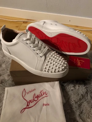 Vita Louboutin ÖPPEN FÖR ALLA BUD - Säljer ett par vita sneakers från Christian Louboutin med ikoniska röda sulor och coola nitar på tån. Skorna är i läder och har klassisk låg profil med vita snören. Perfekta för dig som vill sticka ut med en lyxig och edgy stil.