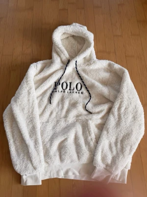 Vit teddyhoodie från Polo Ralph Lauren - Mysig vit hoodie i teddy-material från Polo Ralph Lauren. Stor broderad logga framtill och svarta snören med vita detaljer i huvan. Perfekt för dig som vill ha en stilren och bekväm look med extra fluffig känsla. Ribbade muddar vid ärmslut och nederkant.
