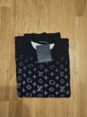 Mörkblå Louis Vuitton stickad tröja - Snygg mörkblå stickad tröja från Louis Vuitton med klassiskt vitt monogrammönster över hela plagget. Tröjan har rund halsringning och ribbade muddar vid ärmslut och nederkant. Perfekt för dig som vill ha en exklusiv och trendig look.