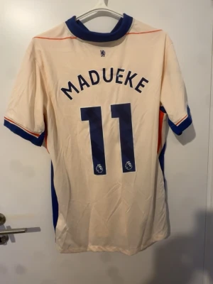 Chelsea Madueke matchtröja nr 11 - Säljer en beige Chelsea FC matchtröja från Nike med Madueke och nummer 11 på ryggen. Tröjan har blå detaljer vid krage och ärmslut, samt klubbmärke och Nike-logga på bröstet. Perfekt för fotbollsfans som vill stötta sitt lag.