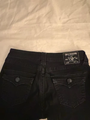 Svarta True Religion Joey jeans - Säljer ett par svarta True Religion Joey jeans med låg midja och flare-modell. Jeansen har snygga fickor med lock och knapp bak. 