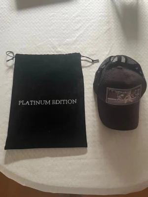 Svart keps med tennisbild - Snygg svart keps med mesh baktill och en unik patch framtill som visar en vintage tennisscen. Kepsen har böjd skärm och justerbar passform. Medföljer svart påse med texten 'PLATINUM EDITION'. Perfekt för dig som gillar sportig och retro stil. Denna keps går ej att köpa i butik eller online.