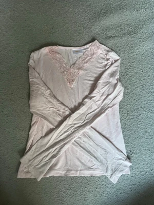 Ljusrosa långärmad loungewear topp - Mysig ljusrosa långärmad topp med v-ringning och spetsdetaljer runt halsen. Tillverkad i mjukt och tunt material som passar perfekt för avslappnade dagar hemma. Spetsen ger en feminin touch och toppen har en enkel, rak passform.