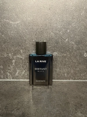 La Rive Distant Wave parfym - Snygg parfymflaska från La Rive i mörkblått glas med svart lock. Flaskan har en stilren, fyrkantig form och texten 'Distant Wave' på framsidan. Perfekt för dig som gillar eleganta och moderna dofter.