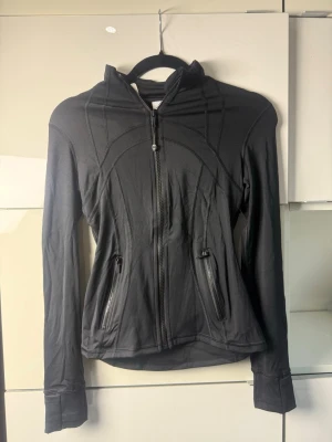 Tröja från Lululemon - Säljer en svart tröja från Lululemon med dragkedja framtill och två fickor med dragkedja. Snygga sömdetaljer och figurnära passform. Perfekt för dig som gillar sportig och stilren look. Materialet känns mjukt och stretchigt.