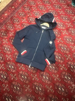Mörkblå hoodie med dragkedja - Snygg mörkblå moncler hoodie med dragkedja och huva. Har vita och röda detaljer på muddar och huva samt en liten patch på ärmen. Tillverkad i mjukt bomullsmaterial och har två fickor framtill. Perfekt för en avslappnad och sportig stil.