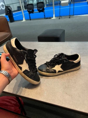 Golden goose svarta 43 - Säljer ett par svarta Golden Goose sneakers med vit stjärndetalj på sidan och vit häl. Skorna har svarta skosnören, något smutsad sula för den klassiska worn looken. Tillverkade i läder och har en låg profil som passar till många stilar. Skicket ser du på bilderna! Priset är inte hugget i sten o det är bara o höra av dig vid minsta fråga eller intresse!😁🙌