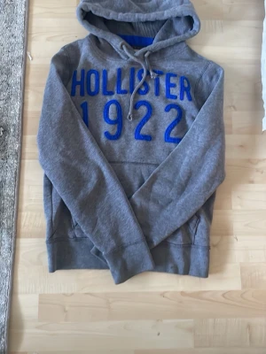 Grå hoodie från Hollister med blå text - Säljer en grå hoodie från Hollister med stor blå text 'HOLLISTER 1922' på bröstet. Hoodien har huva med dragsko och en klassisk känguruficka framtill. Mjuk bomullsblandning och ribbade muddar vid ärmar och nederkant.