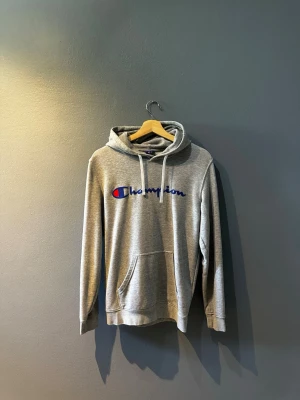 Grå hoodie från Champion - Säljer en grå hoodie från Champion med klassisk logga broderad i blått, vitt och rött på bröstet. Hoodien har huva med vita snören och en stor magficka. Tillverkad i mjukt bomullsmaterial, perfekt för chill dagar eller streetwear-stil.