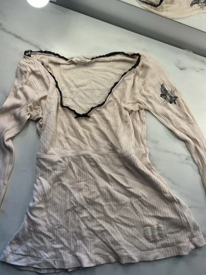 Beige ribbad långärmad topp med spets - Snygg beige ribbad topp med lång ärm och v-ringning dekorerad med svart spetskant. Toppen har ett tryckt motiv på ena ärmen och en liten patch nertill..