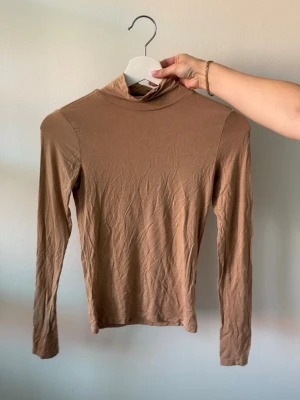 Beige polotopp med lång ärm - Säljer en enkel och stilren beige polotopp med lång ärm. Toppen har en hög krage och är tillverkad i ett mjukt material som känns skönt mot huden. 
