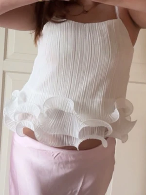 Vit plisserad linnetopp med volang - Säljer en vit linnetopp med tunna axelband och plisserat tyg. Toppen har en unik volangkant nedtill som ger en cool och lekfull look. Perfekt för dig som vill sticka ut lite extra i sommar.