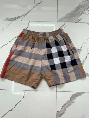 Beige rutiga shorts med snörning - Snygga shorts i beige med klassiskt rutigt mönster i svart, vitt och rött. Shortsen har elastisk midja med snörning och sidofickor. Perfekta för sommaren och ger en trendig look. Sitter som storlek M-L.