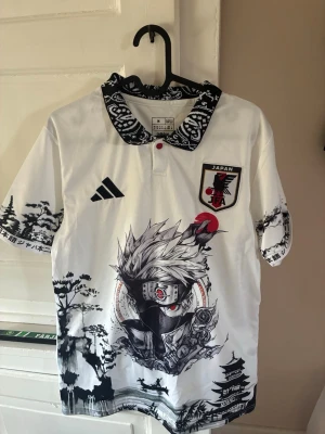 Vit Japan tröja med anime-tryck - Säljer en vit sporttröja med krage och svartvitt mönster, inspirerad av Japan. Framsidan har ett stort anime-tryck med karaktären Kakashi från Naruto, japanska landskap och Japan-emblem på bröstet. Materialet är troligtvis polyester, perfekt för sport eller streetwear.