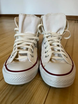 Vita Converse Chuck Taylor All Star - Klassiska vita Converse Chuck Taylor All Star high tops med canvasöverdel, vita snören och gummisula. Skorna har den ikoniska röda linjen runt sulan och Converse-loggan på sidan. Perfekta för en avslappnad och stilren look.